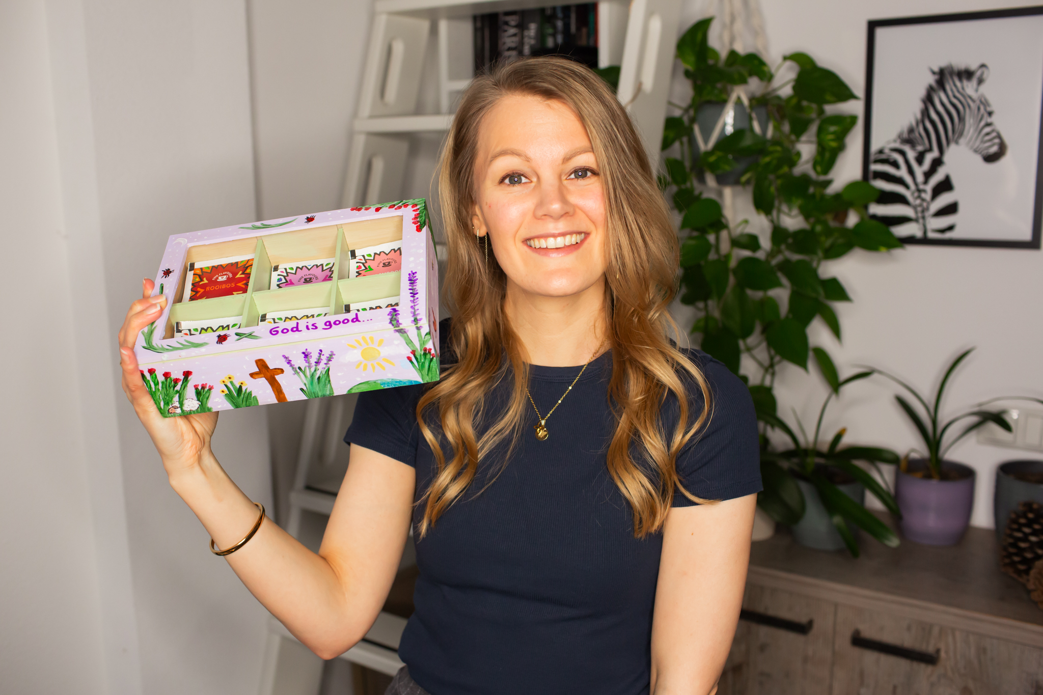 Deborah lacht en houdt een beschilderde theedoos in haar handen gevuld met kleurrijke theezakjes